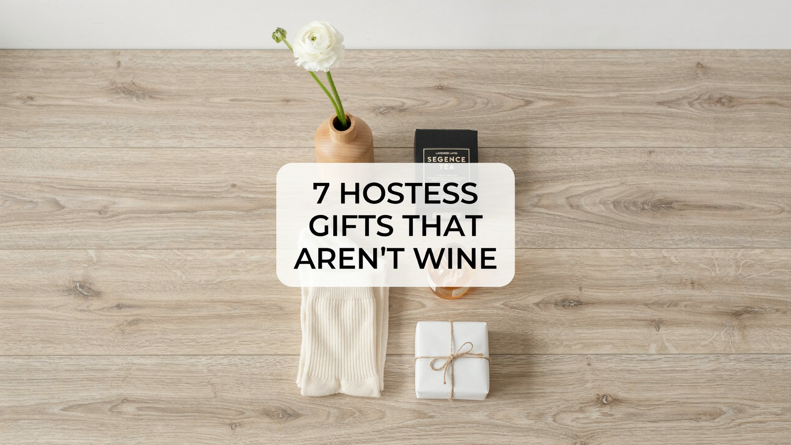 Hostess Gifts: pale oak, shot in bright cool daylight. Running down the frame: a slim wood bud vase with a single white stem, a folded pair of cream cashmere socks, a small box of tea