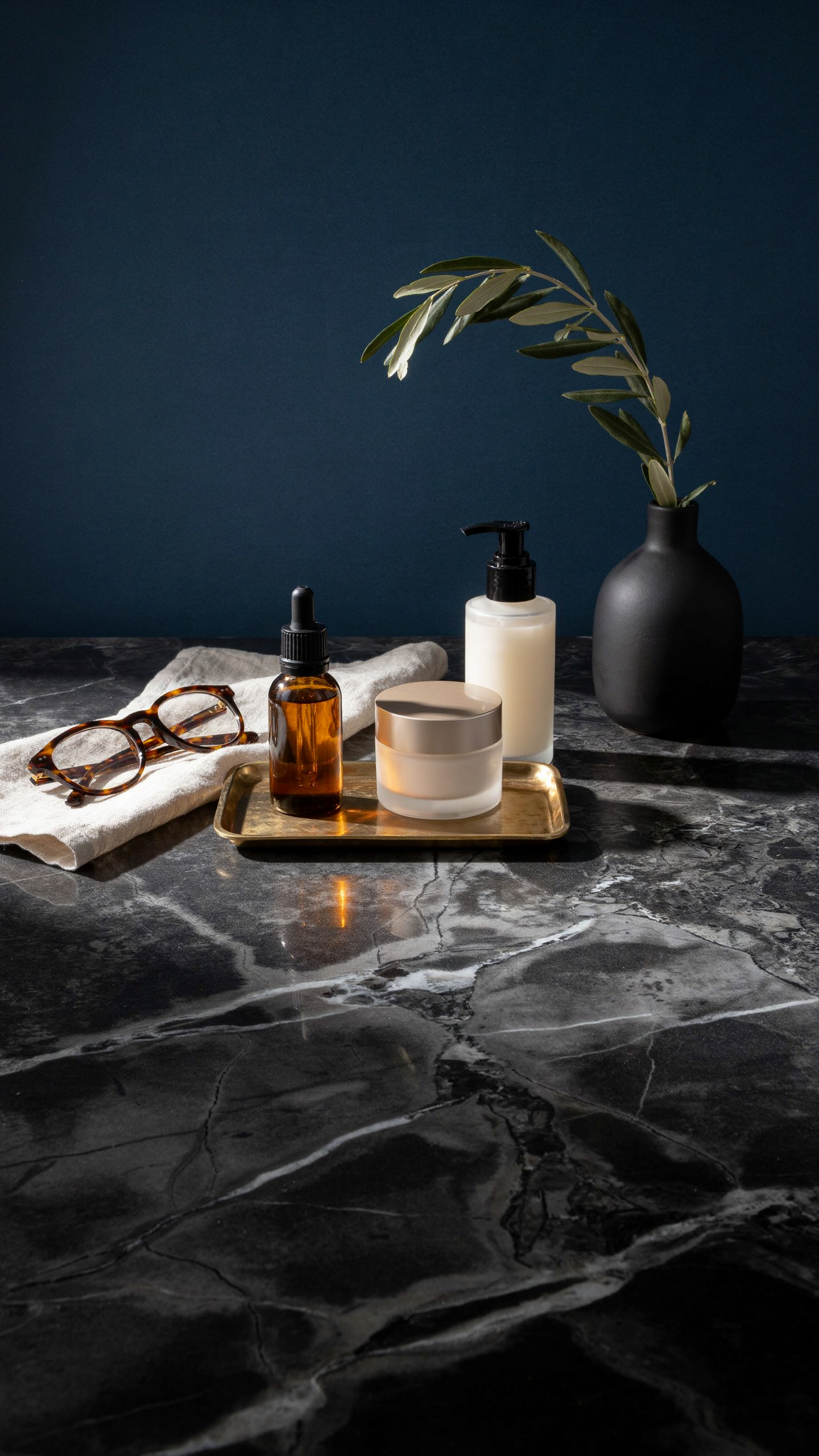 Amber skincare bottle, cream moisturizer, cleanser, reading glasses, and ivory towel arranged on a gold tray over black marble
