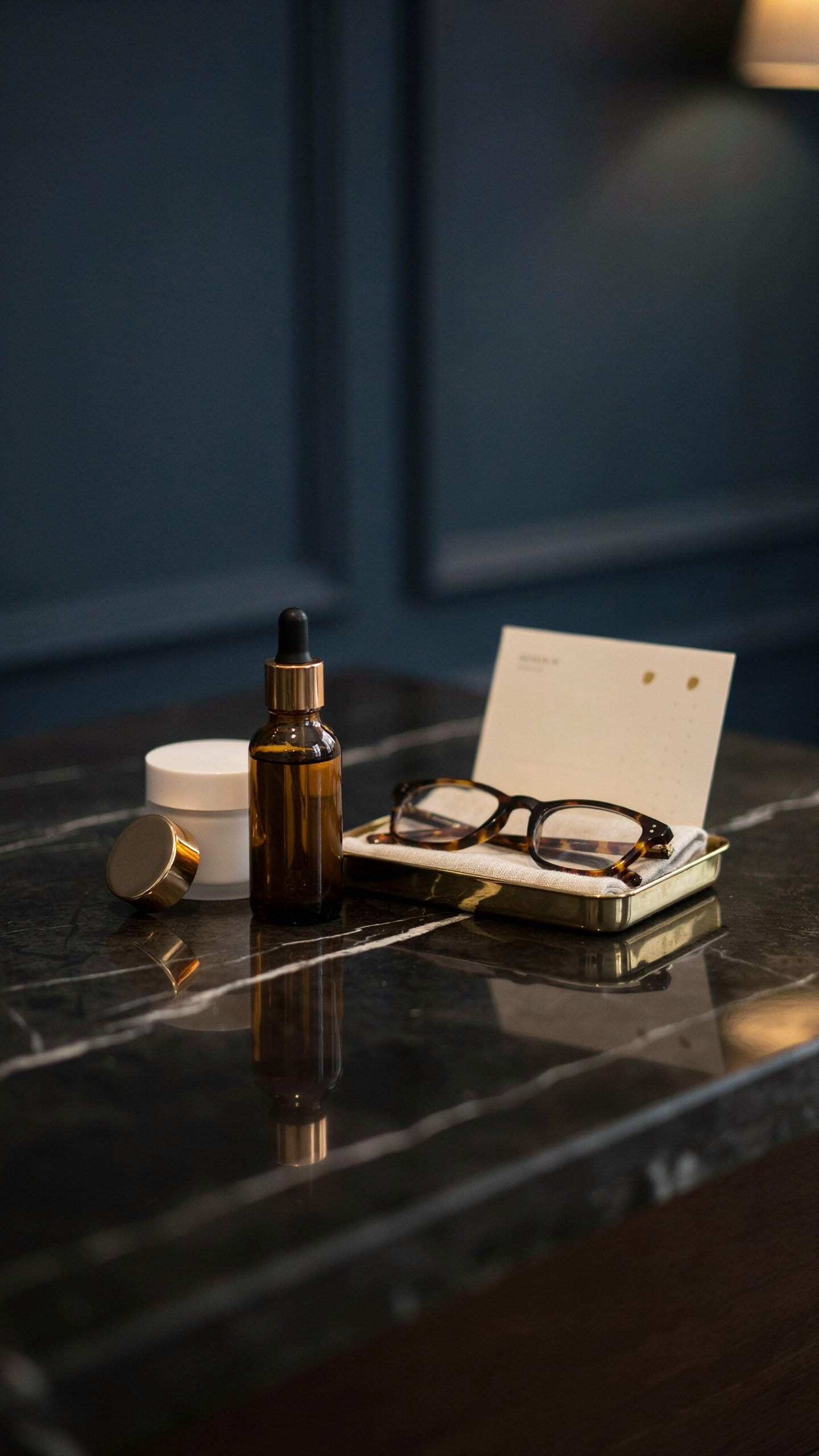 Amber skincare bottle, cream moisturizer, reading glasses, and a small retinol calendar arranged on a black marble vanity